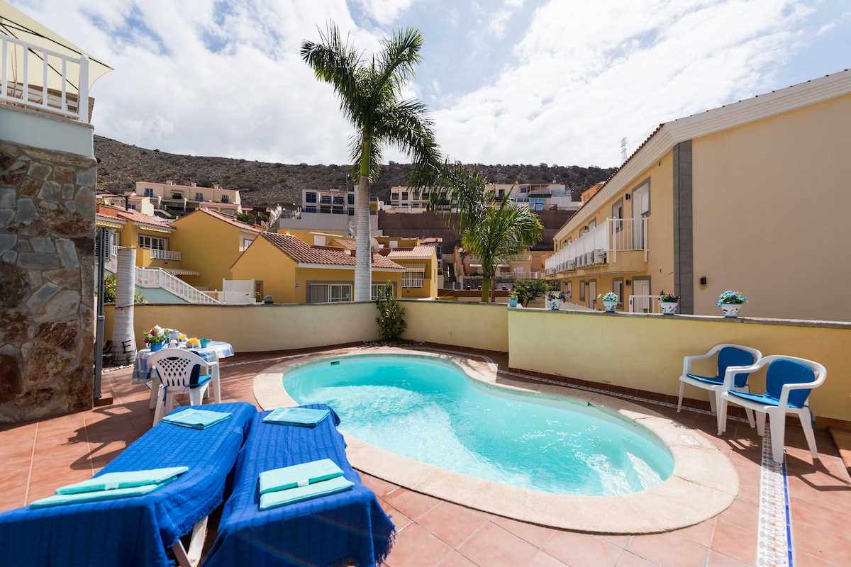 A private saltwater pool is surrounded by a sun terrace, featuring two lounge chairs with blue cushions. A few white patio chairs are placed nearby. Lush green foliage and nearby buildings create a cozy atmosphere, with a hillside visible in the background.