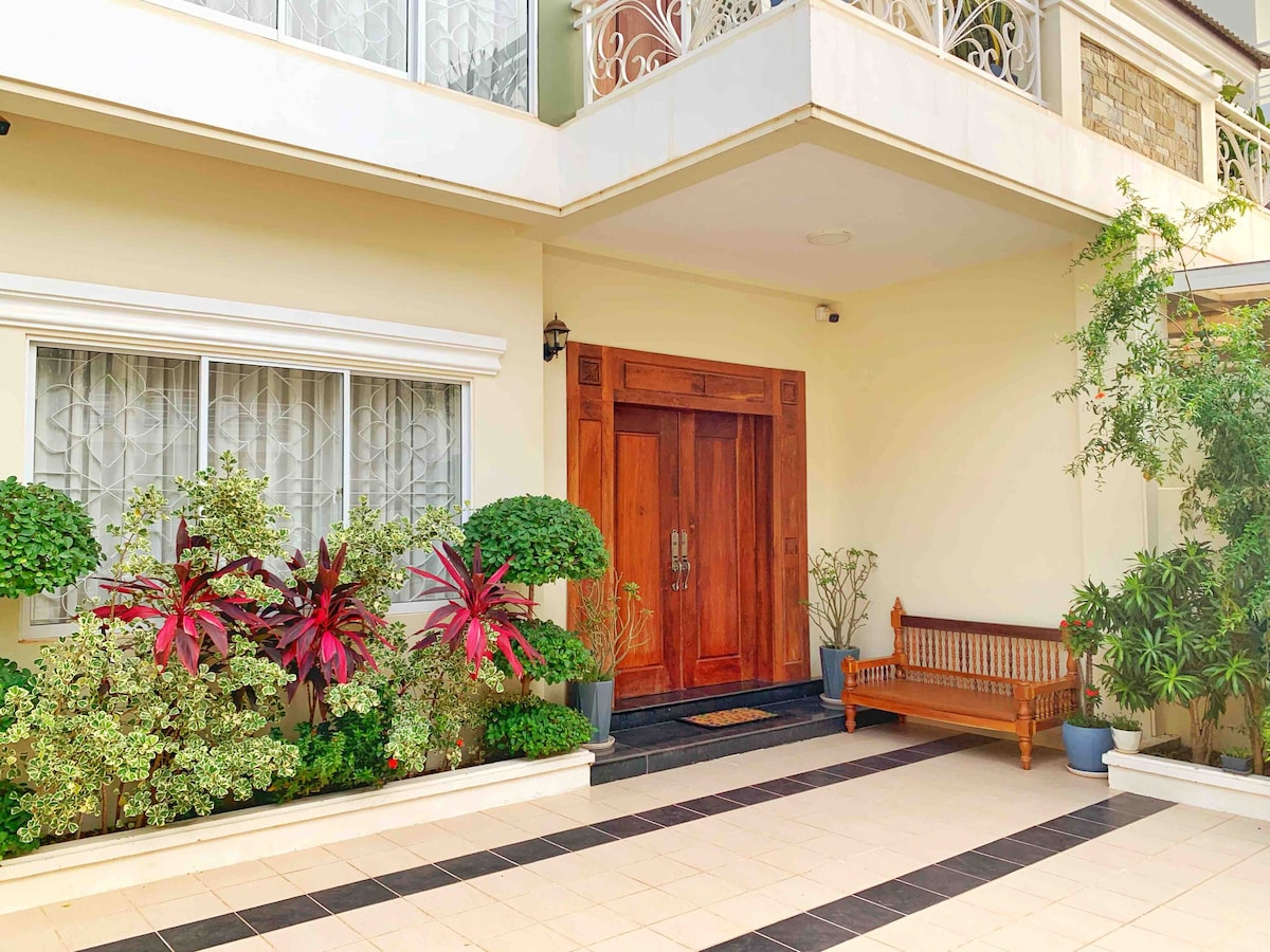 The entrance features a large wooden double door framed by lush greenery, including potted plants and low shrubs. A wooden bench is positioned to the side, providing a welcoming space to relax. Expansive windows allow natural light to brighten the area, adding to its inviting ambiance.