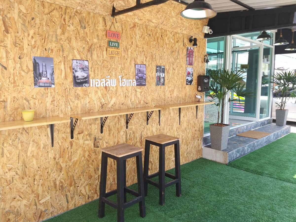 An open co-living area is depicted, featuring a rustic wooden wall with decorative posters. Two high stools sit at a long wooden bar, complemented by small potted plants. Natural light enters through large windows, enhancing the inviting atmosphere of the space.