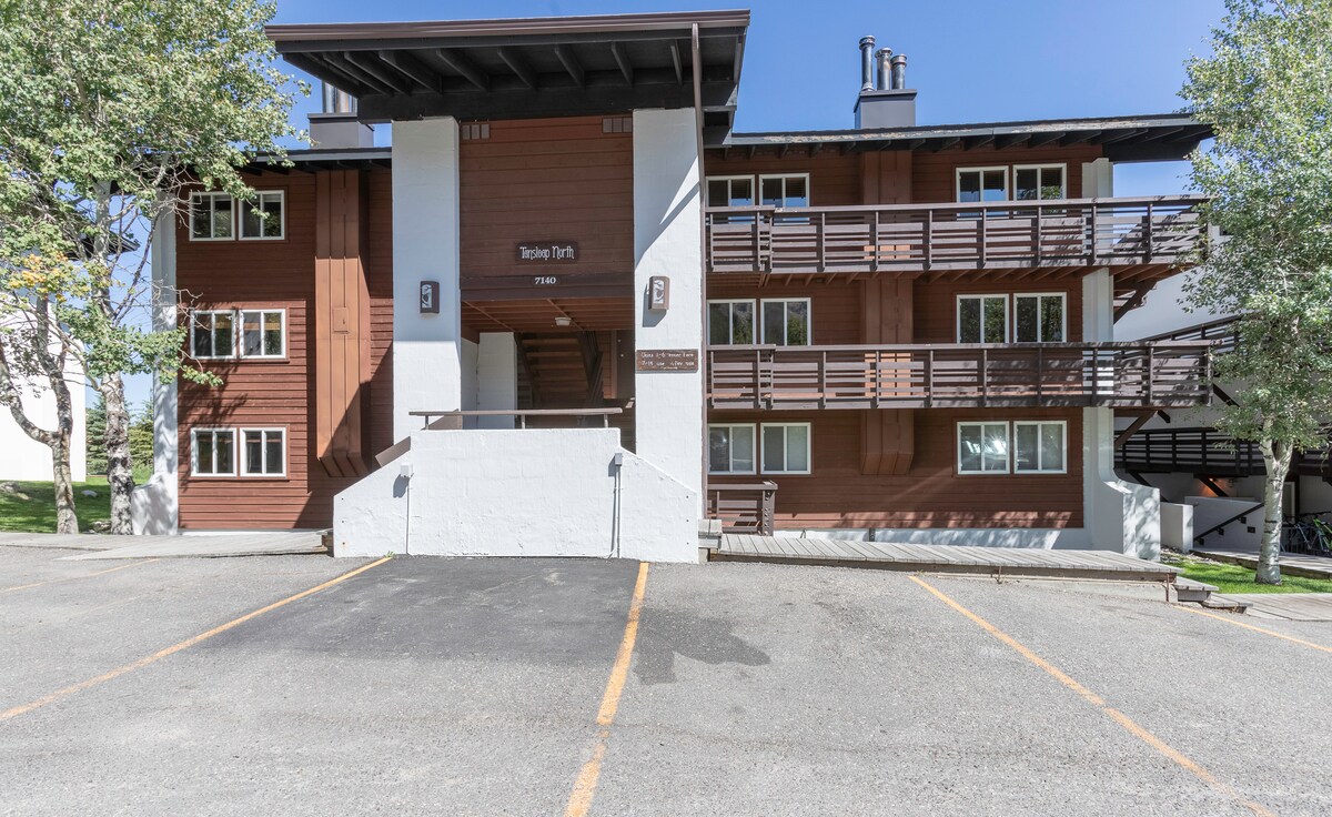 The exterior of a multi-unit condo building features a combination of dark wood and light stucco elements. A set of stairs leads to the entrance, while multiple balconies are visible on the upper levels. A spacious parking area is provided in front.