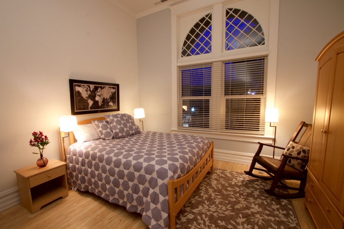 A well-appointed bedroom features a spacious bed with a patterned quilt against light blue walls. A wooden rocking chair is positioned beside a nightstand with a small vase of flowers. Natural light filters through large windows with decorative grille patterns, creating a serene atmosphere.