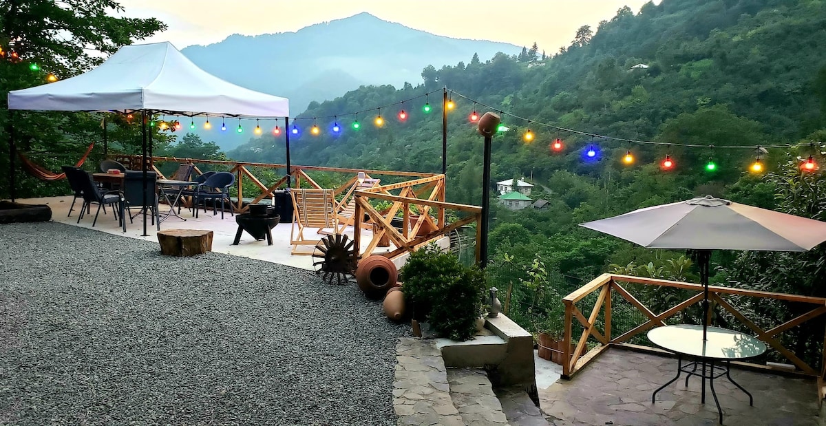 An outdoor patio area is shown, featuring string lights that illuminate the space. A white canopy provides shade over a wooden table and chairs, while a hammock is positioned nearby. Scenic mountains create a tranquil backdrop, enhancing the relaxed atmosphere.