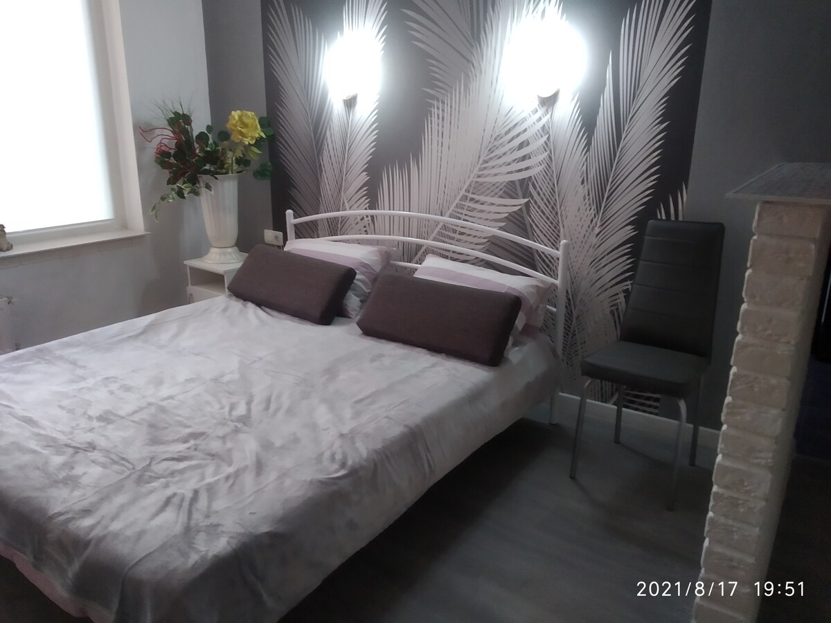 A spacious bed is showcased in the room, adorned with soft gray bedding and accent pillows. A light-colored wall features a tropical plant design, complemented by strategically placed wall sconces. A single modern chair sits nearby, adding functionality to the space.
