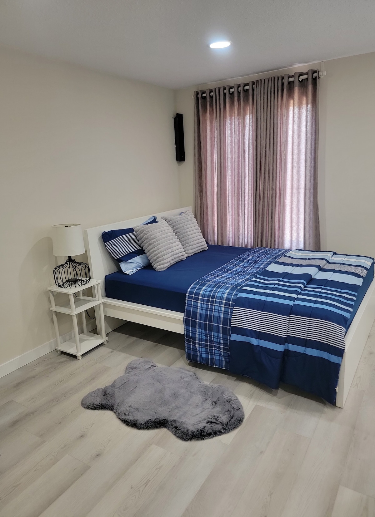 A neatly arranged sleeping area features a spacious bed dressed in a blue and white striped duvet and complemented by two decorative pillows. Natural light enters through window coverings, and a small bedside lamp rests on a white nightstand beside the bed. A soft area rug is laid on the floor.
