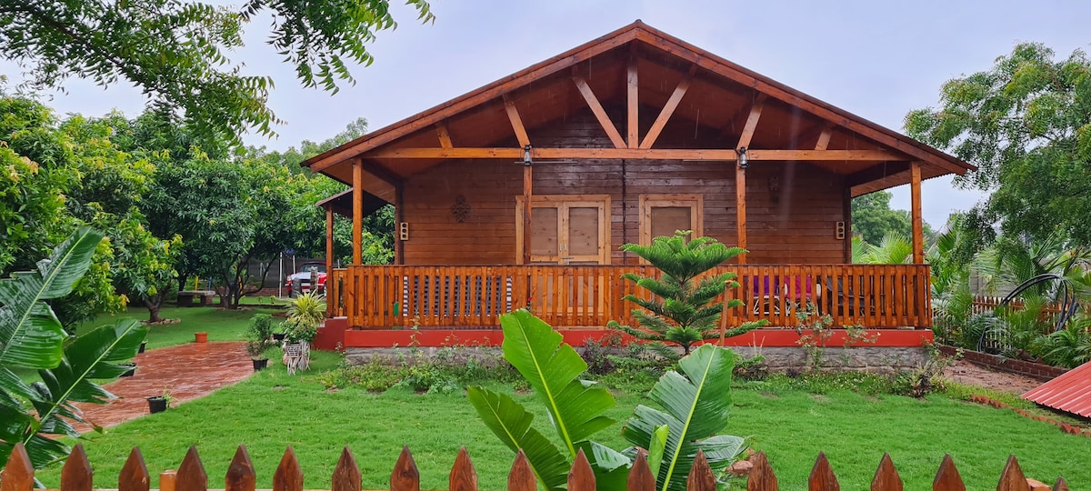 A rustic wooden cottage is set amidst lush green gardens, showcasing a spacious front porch with wooden railings. Tropical plants and trees surround the area, offering a serene atmosphere. The cottage's symmetrical design features large doors, enhancing its charming appeal.