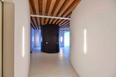 A modern corridor is presented, featuring wooden ceiling beams that contrast with the smooth white walls. Soft lighting is provided by slender wall sconces on either side, creating a serene ambiance. A circular black structure is visible at the end of the hallway, adding unique architectural interest.