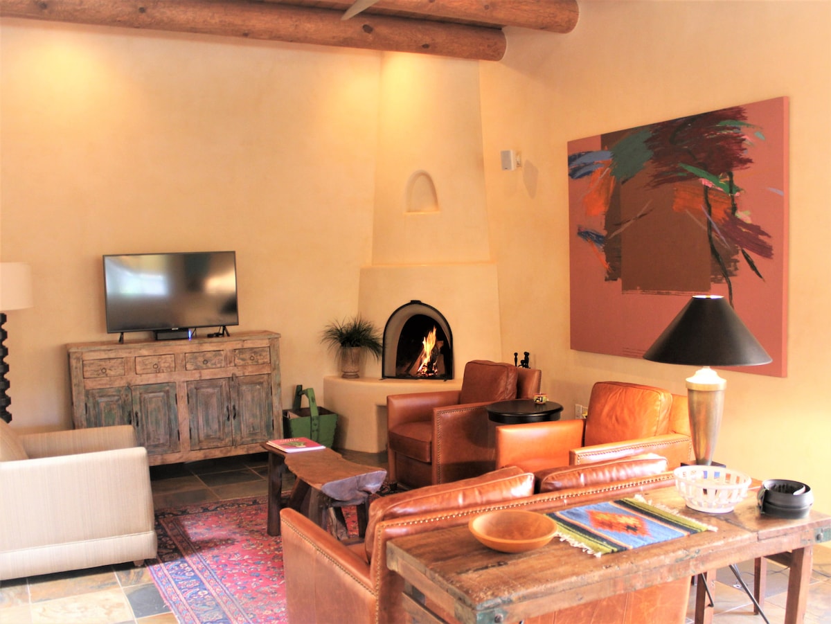 The living area is furnished with leather seating and a wooden coffee table. A Kiva fireplace provides warmth and is accompanied by a flat-screen TV. Colorful artwork decorates the wall, and soothing natural light illuminates the space through skylights.