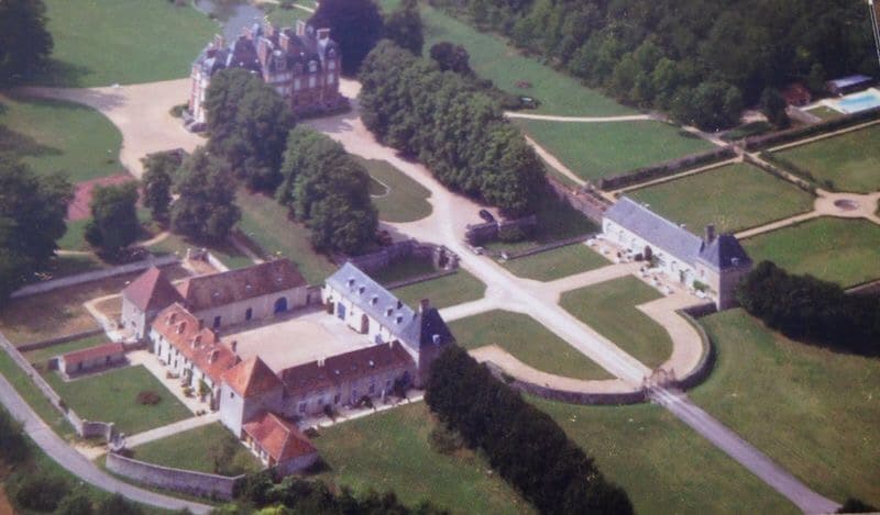 An aerial view captures the expansive grounds of Chateau La Trousse, featuring a grand main building surrounded by lush green lawns and mature trees. Pathways, residential buildings, and a swimming pool are visible, illustrating the spacious layout of the estate.