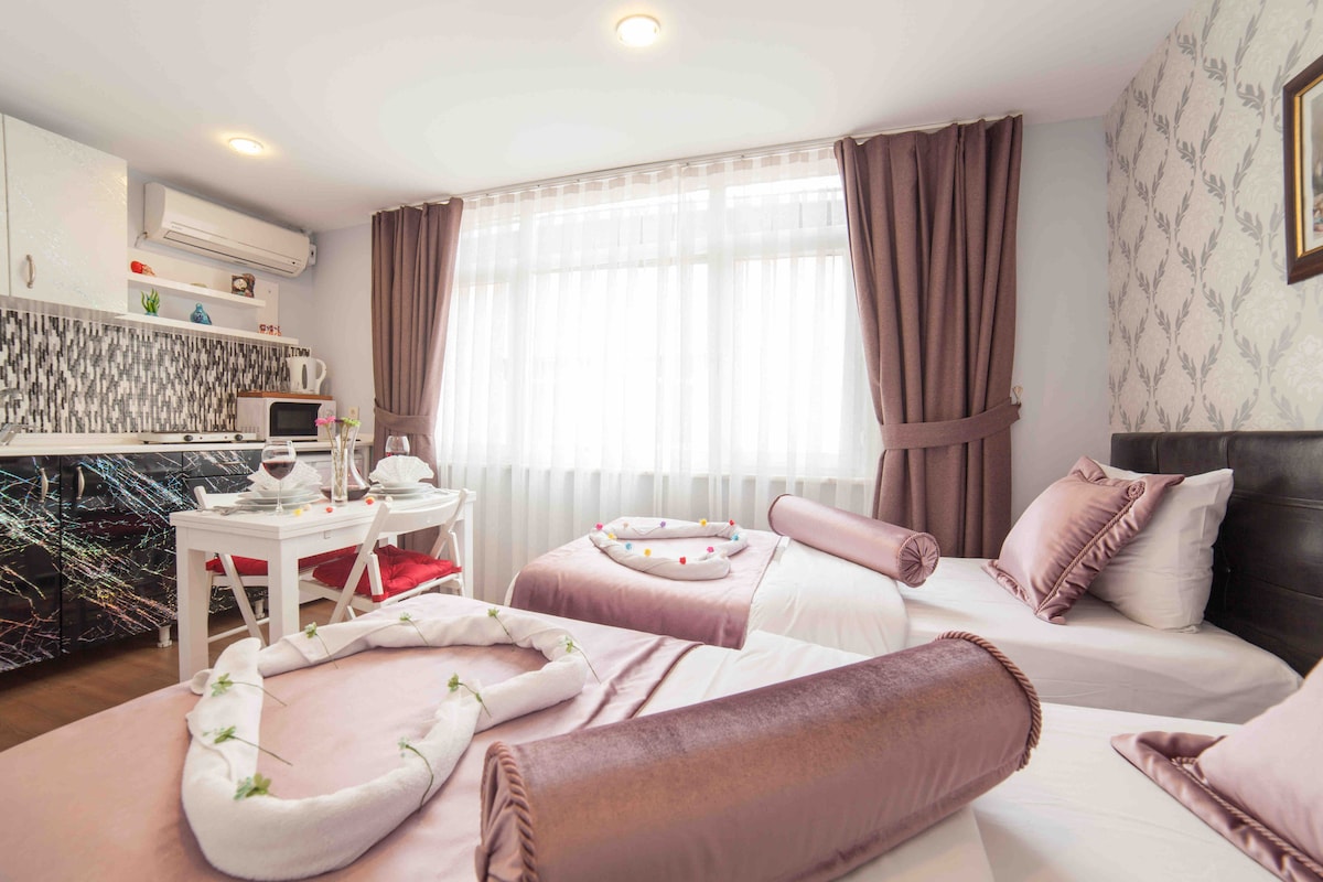 A bright room features two spacious beds adorned with soft pink bedding and decorative bolsters. A dining area with a white table and chairs is visible next to a kitchenette, which includes modern appliances. Natural light floods the space through large windows covered by sheer curtains.