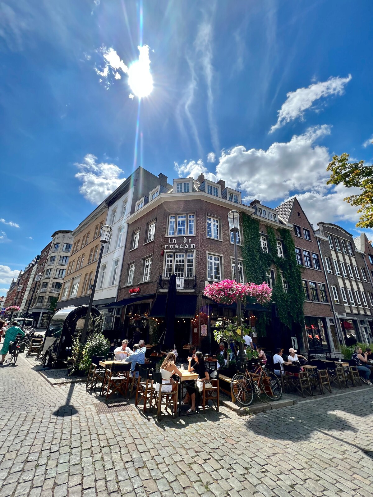 A lively street scene captures a historic building adorned with blooming pink flowers. Outdoor seating is filled with patrons enjoying their meals. Cobblestone paths are visible, along with bicycles parked nearby. The sky is bright, showcasing a few fluffy clouds.