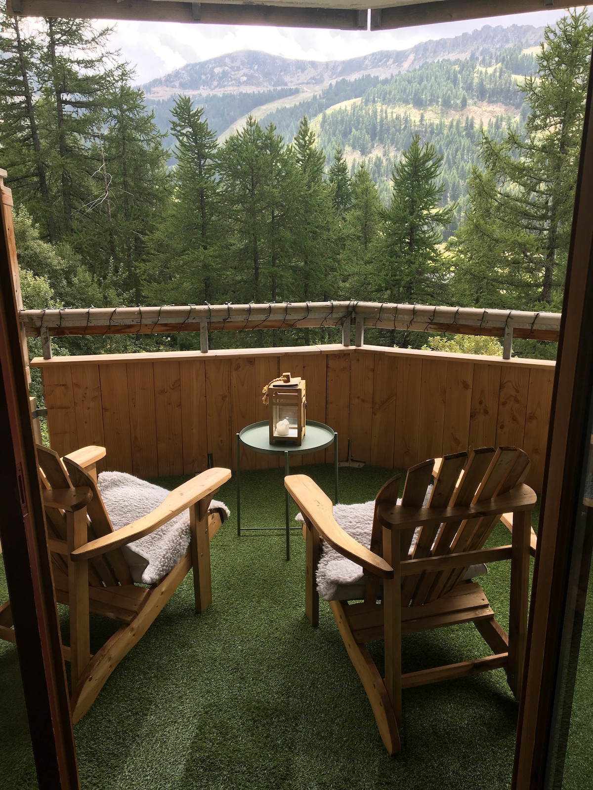 An inviting balcony features two wooden chairs paired with a small round table. Lush green artificial grass covers the floor, while a lantern sits atop the table. The surrounding trees and mountains create a serene backdrop, enhancing the peaceful ambiance.