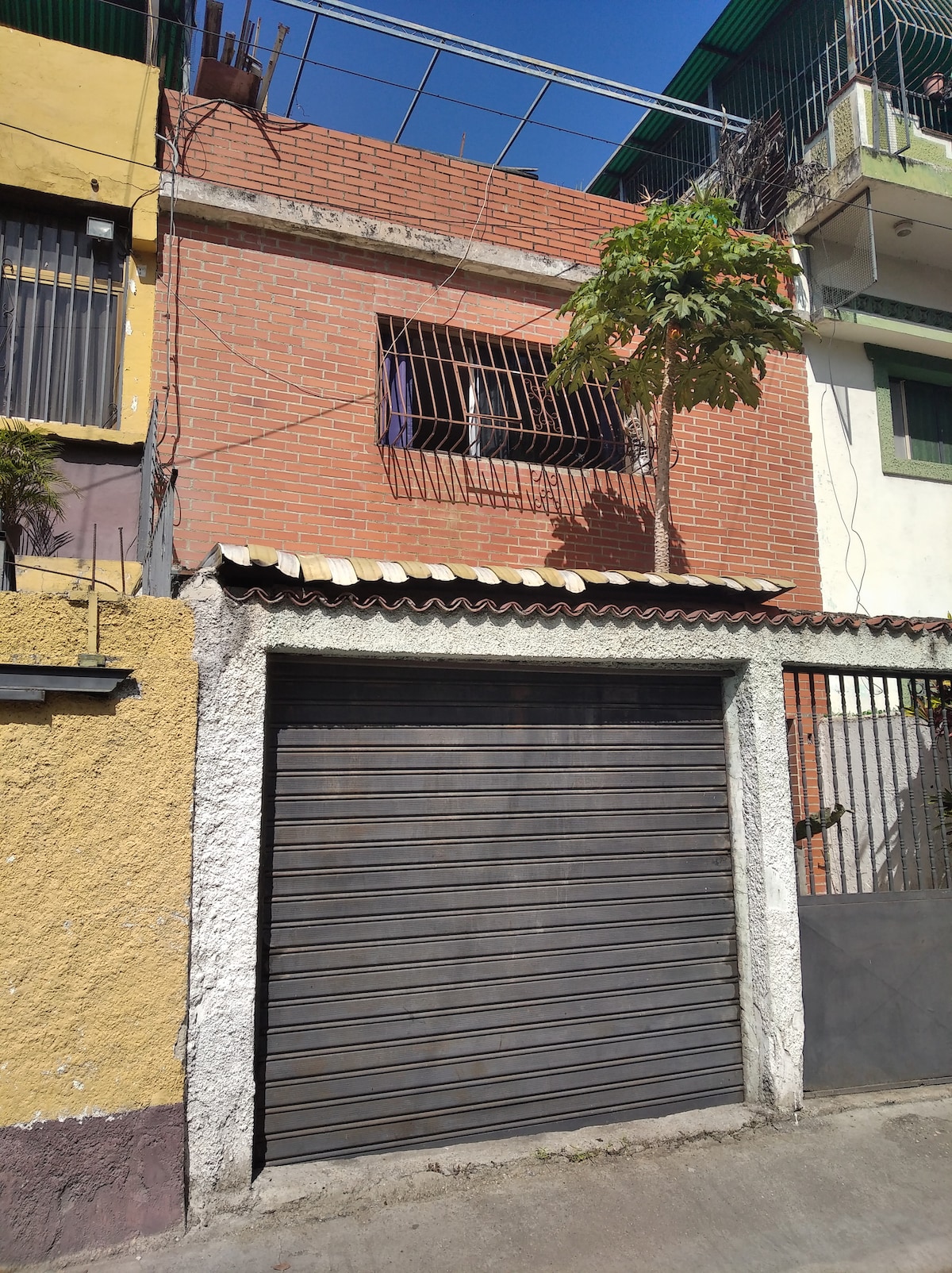 The exterior of a two-story building is visible, featuring a gray garage door at the bottom. Above the garage, a window with protective bars is framed by a red brick wall, and a small tree is placed nearby. Vibrant yellow and green walls of adjacent structures provide contrast.