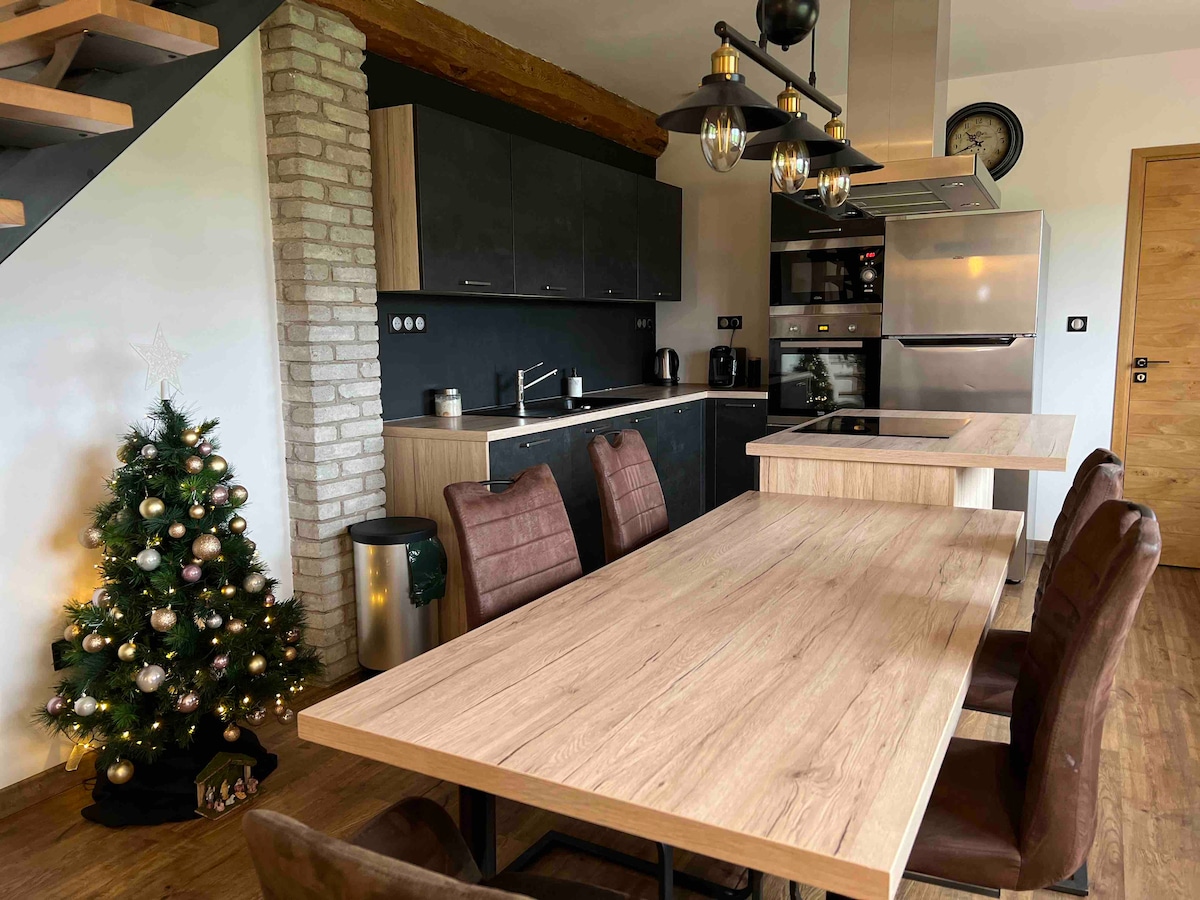 An open kitchen and dining area features modern appliances, including a stove and refrigerator. A wooden dining table is surrounded by four dark chairs. A small Christmas tree decorated with ornaments adds a festive touch to the space, complemented by rustic wooden beams on the ceiling.