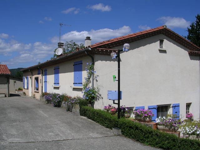 A light-colored building with blue shutters is visible, surrounded by greenery and potted flowers. A gravel path leads to the entrance, and several outdoor light fixtures are positioned along the path. Sky and clouds provide a pleasant backdrop.