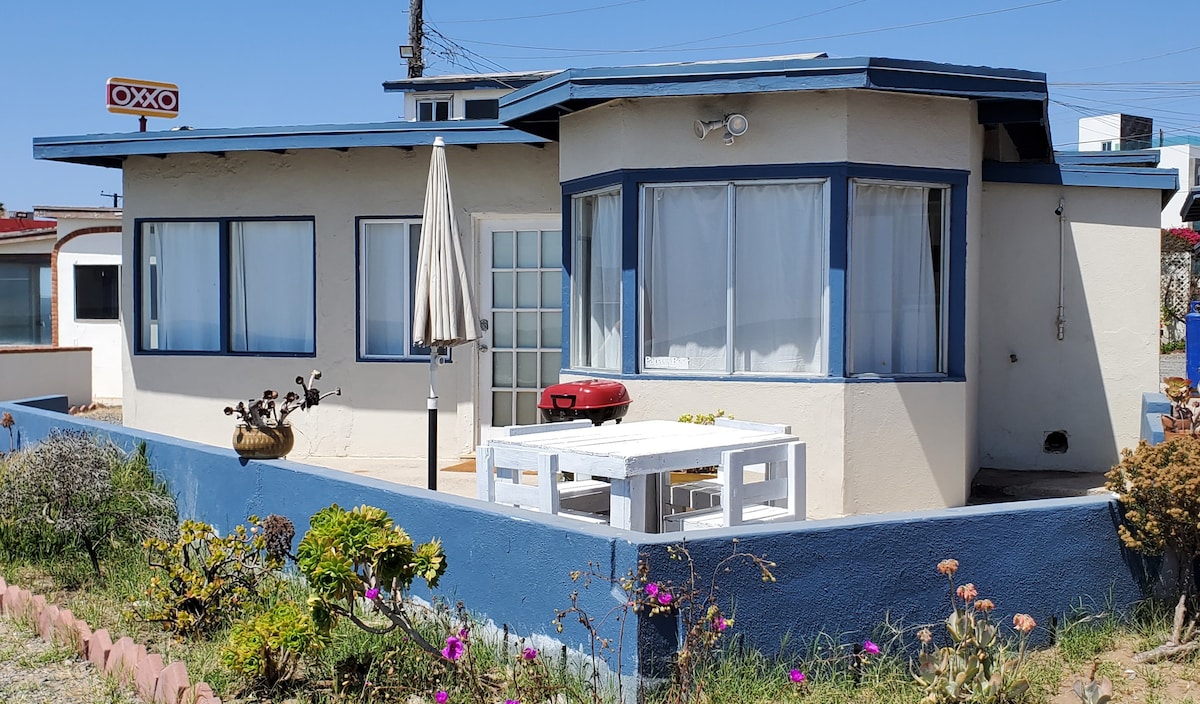 The exterior of the house is showcased, featuring light-colored walls and blue trim. A patio with a table and chairs is visible in front, with an umbrella providing shade. Vibrant flowering plants surround the area, enhancing the coastal charm.