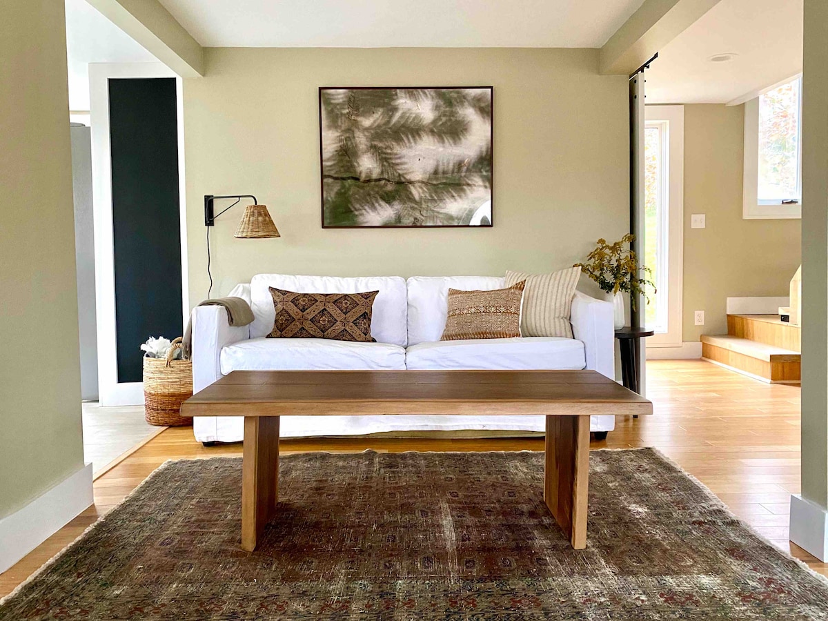 A light-filled living area features a white sofa with decorative cushions, positioned opposite a simple wooden coffee table. A patterned area rug anchors the space, while a framed botanical print adorns the wall. A tall plant adds a touch of greenery near one corner.