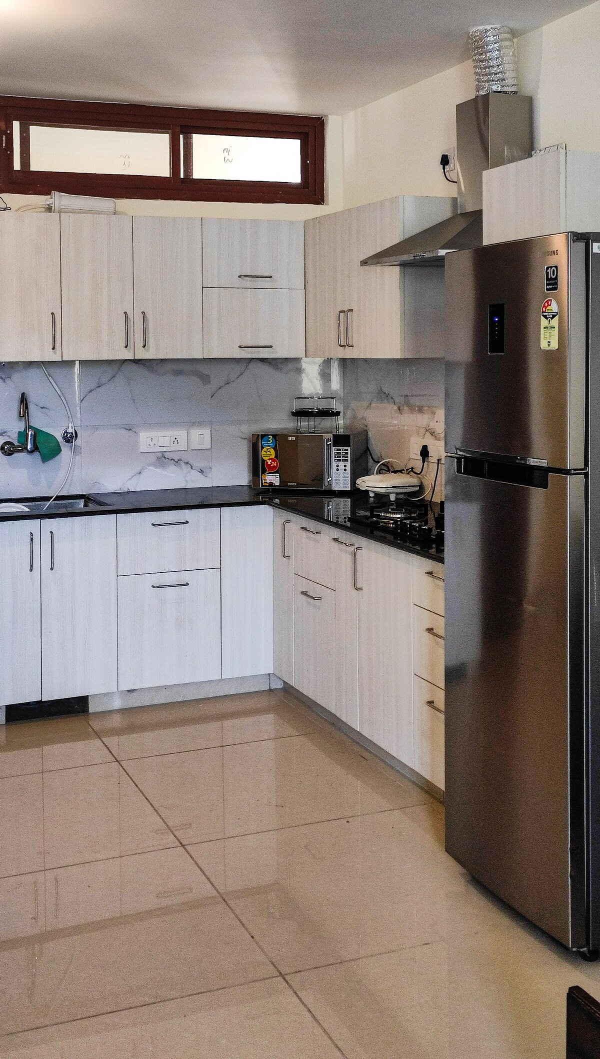 A modern kitchen featuring light wood cabinetry and a sleek stainless steel refrigerator. Equipped with a gas stove and essential appliances, the space is designed with ample storage, complemented by polished floor tiles that reflect natural light.