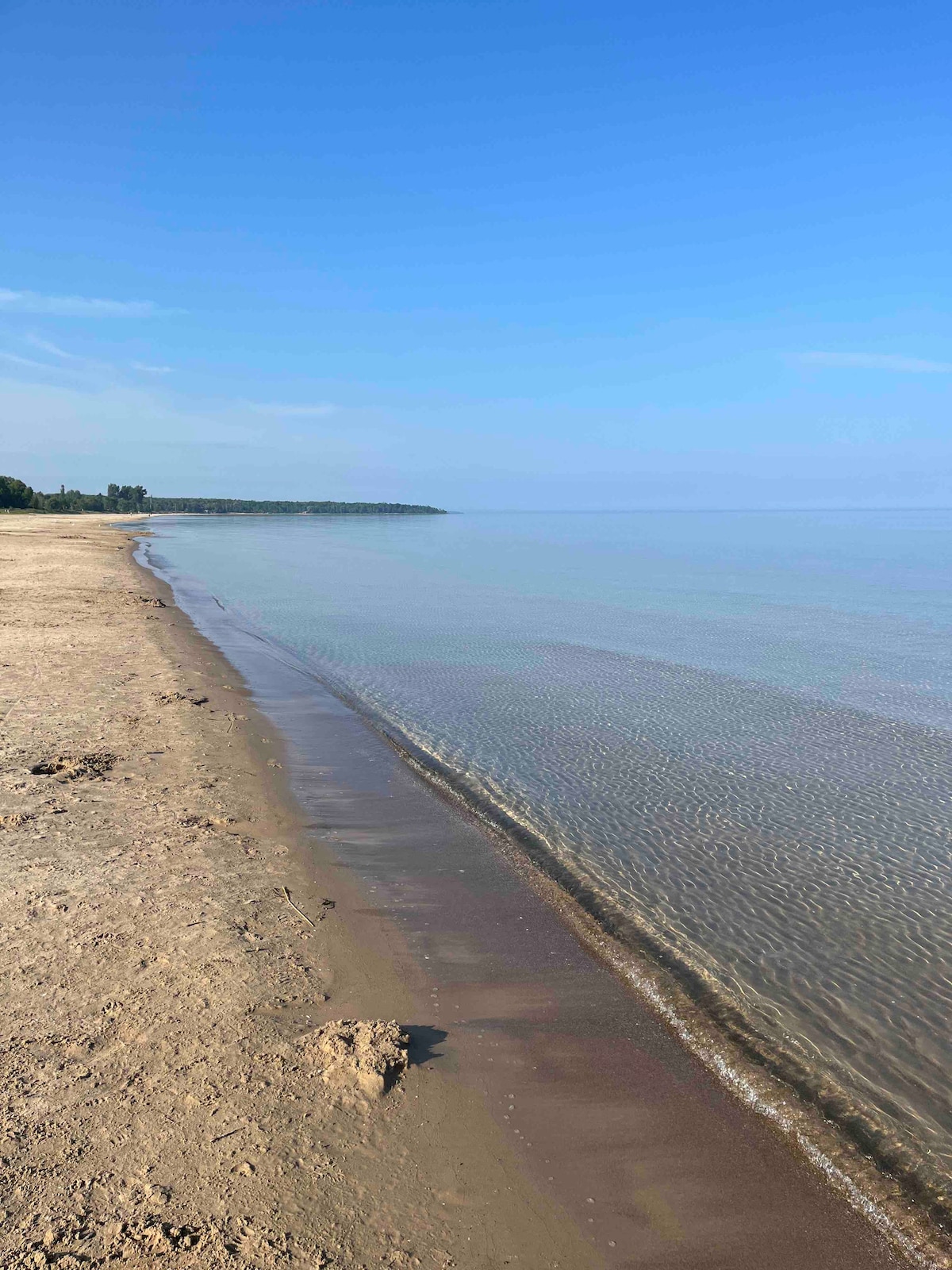 A serene view of a sandy beach stretches along a calm body of water. The shoreline gently curves, with soft waves lapping at the sand. Clear blue skies reflect on the water, creating a peaceful atmosphere.