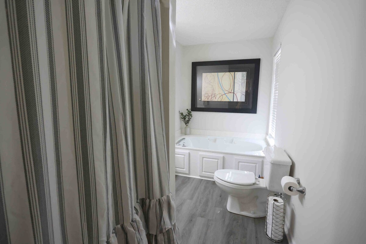 A well-lit bathroom features a large soaking tub and a privacy shower curtain. A framed artwork is mounted on the wall above the tub, enhancing the space. The room includes a toilet, a towel holder, and a window allowing natural light.