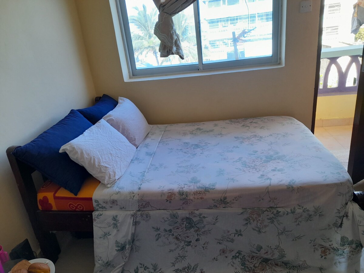 A bright sleeping area features a spacious bed with a floral bedspread and two sets of pillows in blue and white. Natural light flows through a large window, revealing glimpses of the outdoors and providing a serene ambiance.