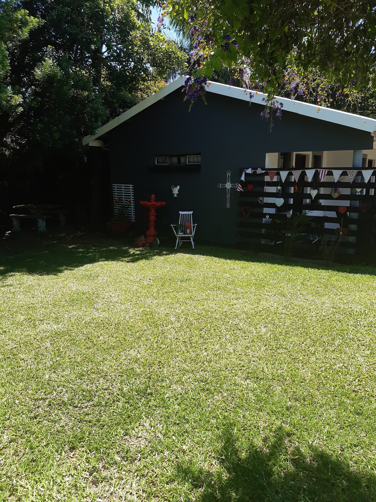 A well-maintained garden area is seen, featuring a lush green lawn. A simple wooden chair is positioned on the grass, and decorative elements are visible on the nearby wall. A small shaded area with trees adds to the tranquil setting, contributing to a serene outdoor atmosphere.