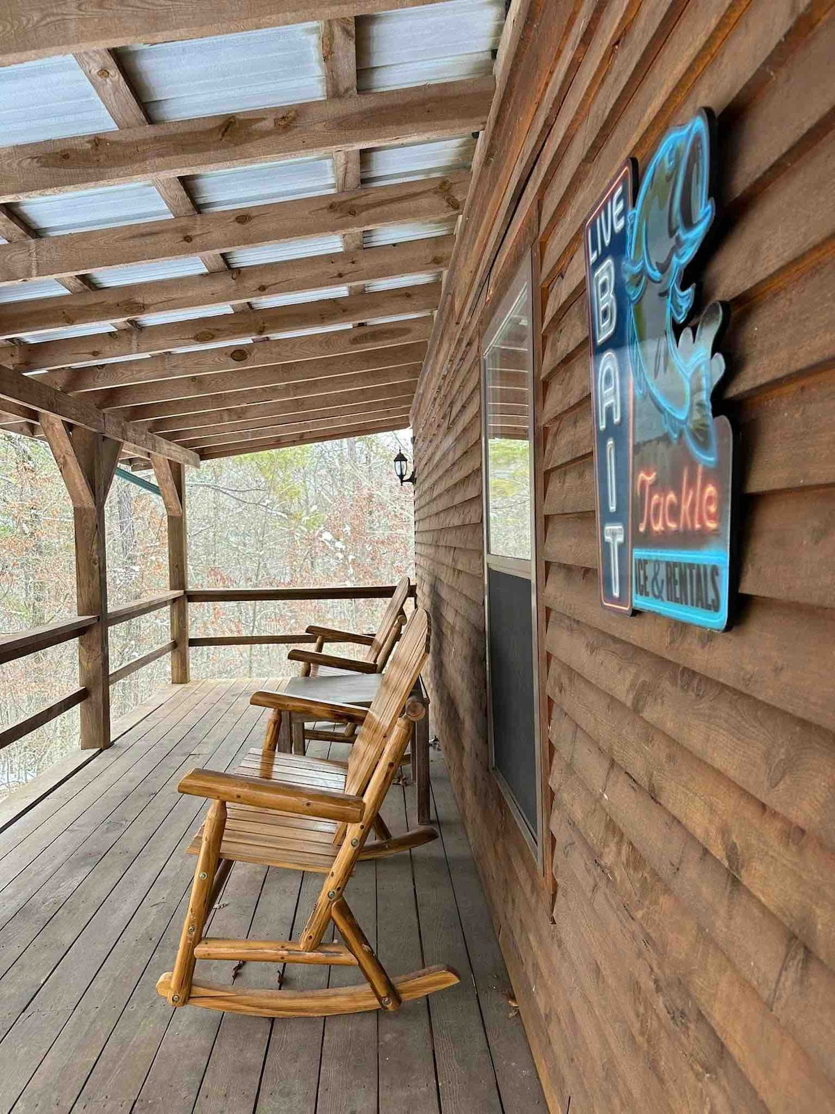 A wooden porch is depicted, featuring several rocking chairs arranged along the railing. A brightly colored sign labeled 'LIVE BAIT' adds a quaint touch to the rustic atmosphere. Natural surroundings are visible in the background, enhancing the cabin's connection to nature.