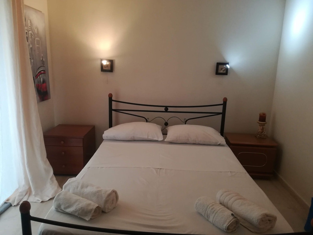 A spacious bed is framed by two bedside tables, each featuring a wall-mounted light fixture. Soft towels are neatly arranged at the foot of the bed. Natural light filters in through a sheer curtain, illuminating the room's neutral walls and offering a relaxed atmosphere.