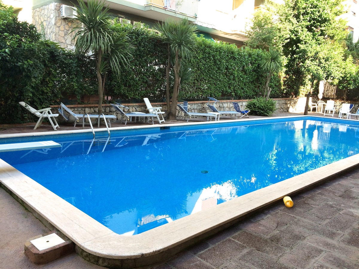 A large swimming pool is surrounded by lush greenery, creating a serene atmosphere. Sun loungers are positioned along the poolside, providing ample space for relaxation. The clear, inviting water reflects the sunlight and nearby plants, enhancing the tranquil environment.