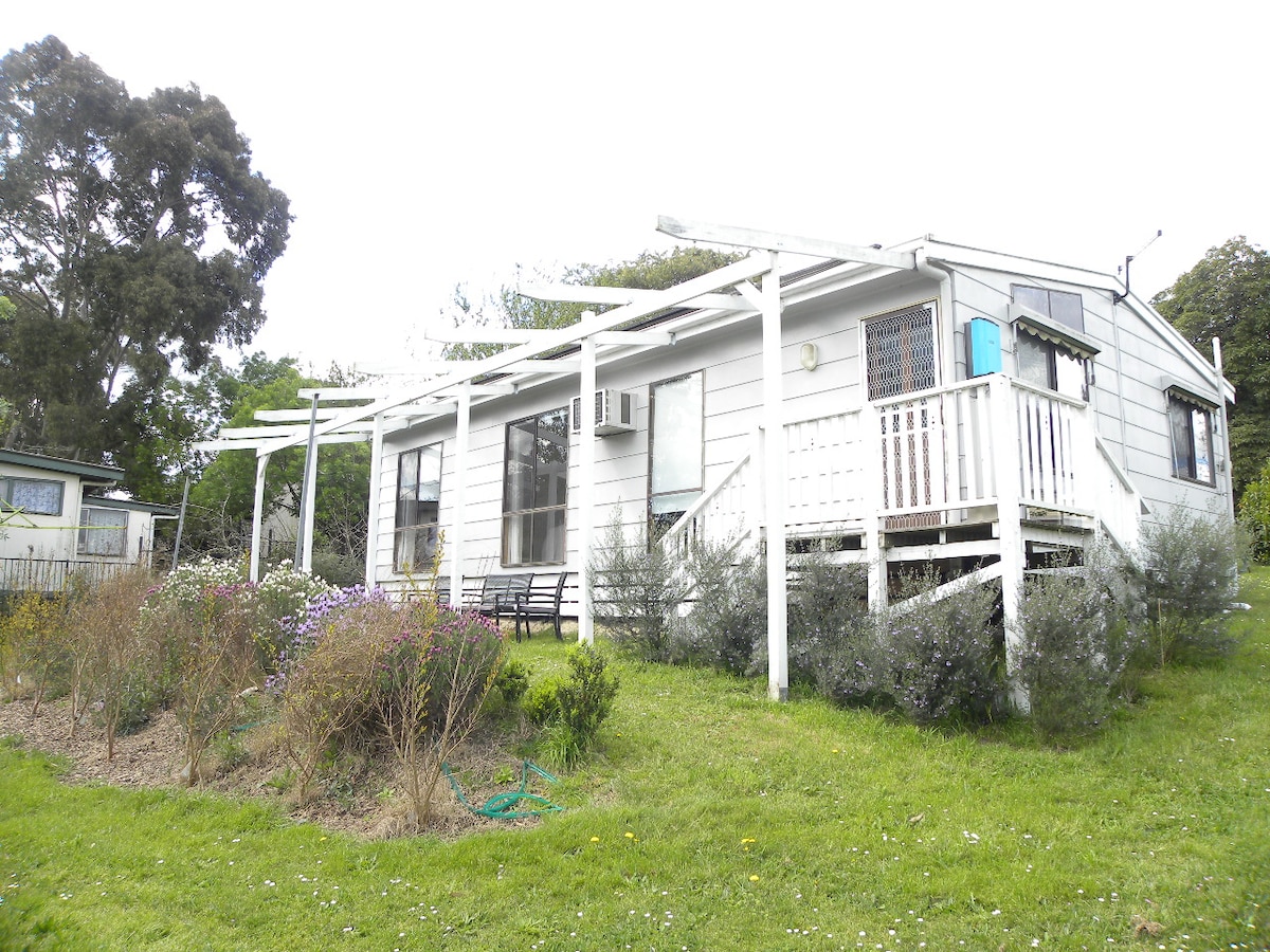 A quaint house features a light exterior and an elevated porch area, surrounded by neatly maintained gardens and pathways. Outdoor seating is visible on the porch, and several shades provide shelter. A serene green lawn enhances the overall charm of the setting.