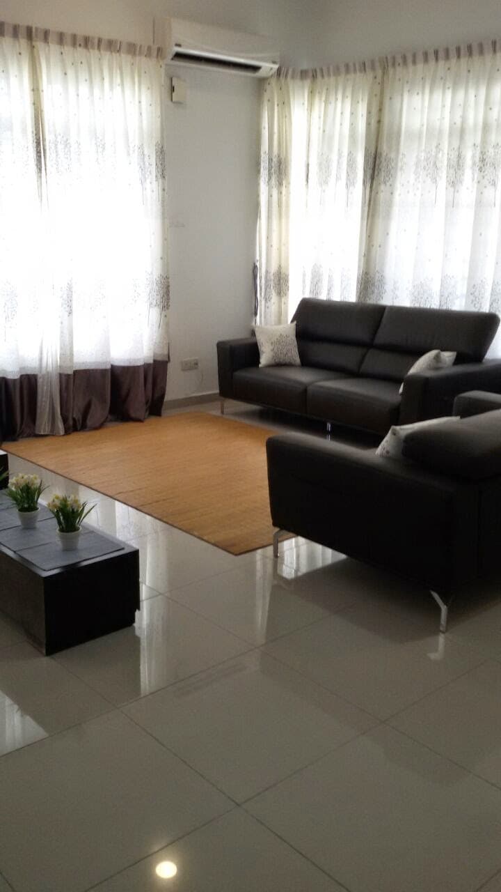 A well-lit living area is furnished with a sleek black sofa, complemented by decorative cushions. A simple rectangular coffee table sits on a light-colored rug. Natural light filters through sheer curtains, creating a bright and welcoming ambiance.