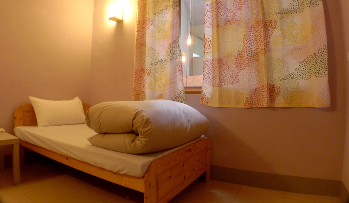 A single bed with a wooden frame is positioned against a pastel pink wall. Two fluffy pillows are stacked on the bed. Colorful, patterned curtains frame a window, allowing natural light to illuminate the room.