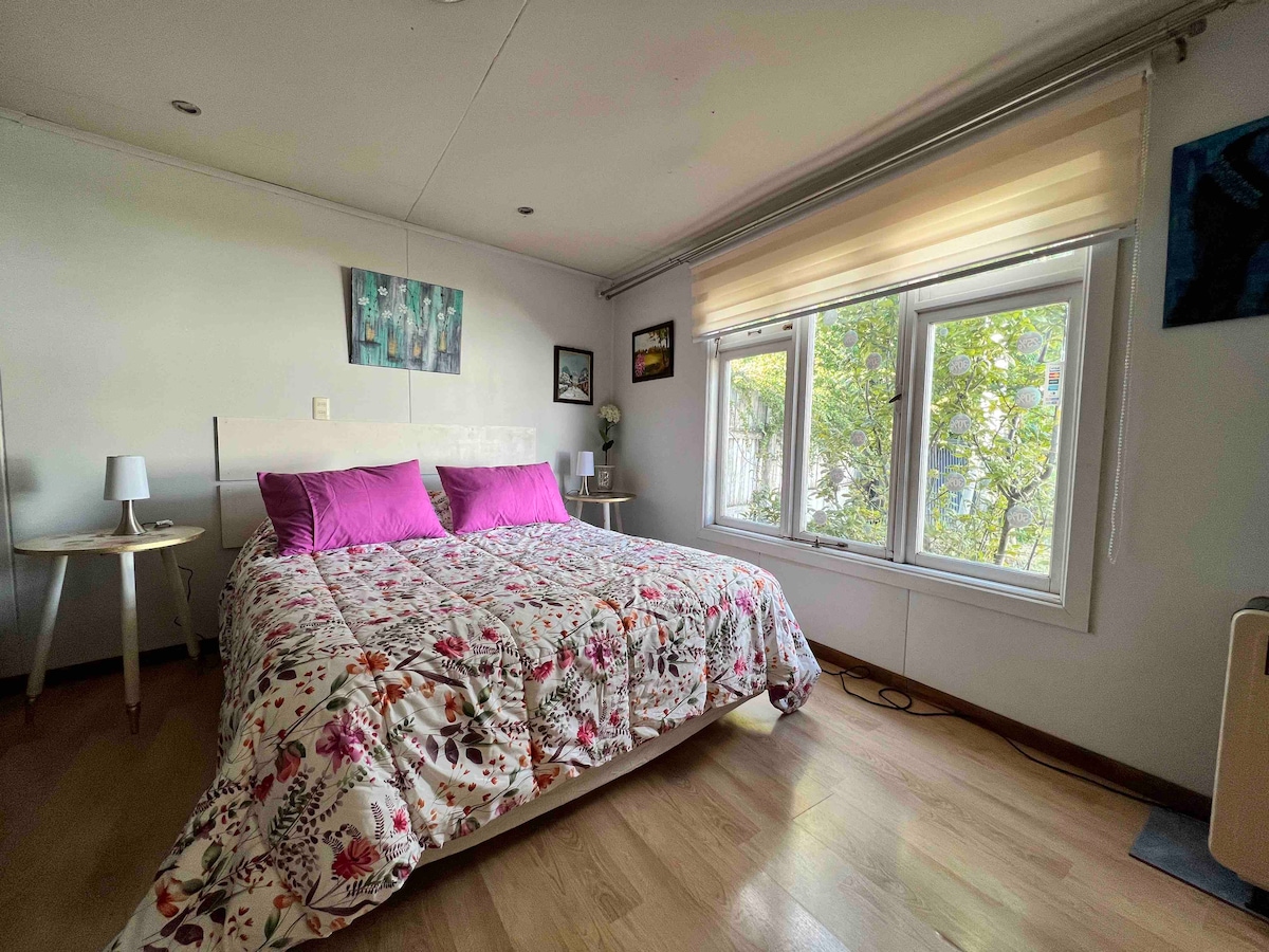 A spacious bedroom is presented with a floral-patterned bedspread and two purple pillows on the bed. Natural light filters through large windows adorned with light, sheer curtains. Two bedside tables with lamps are positioned beside the bed, enhancing the room's functionality.