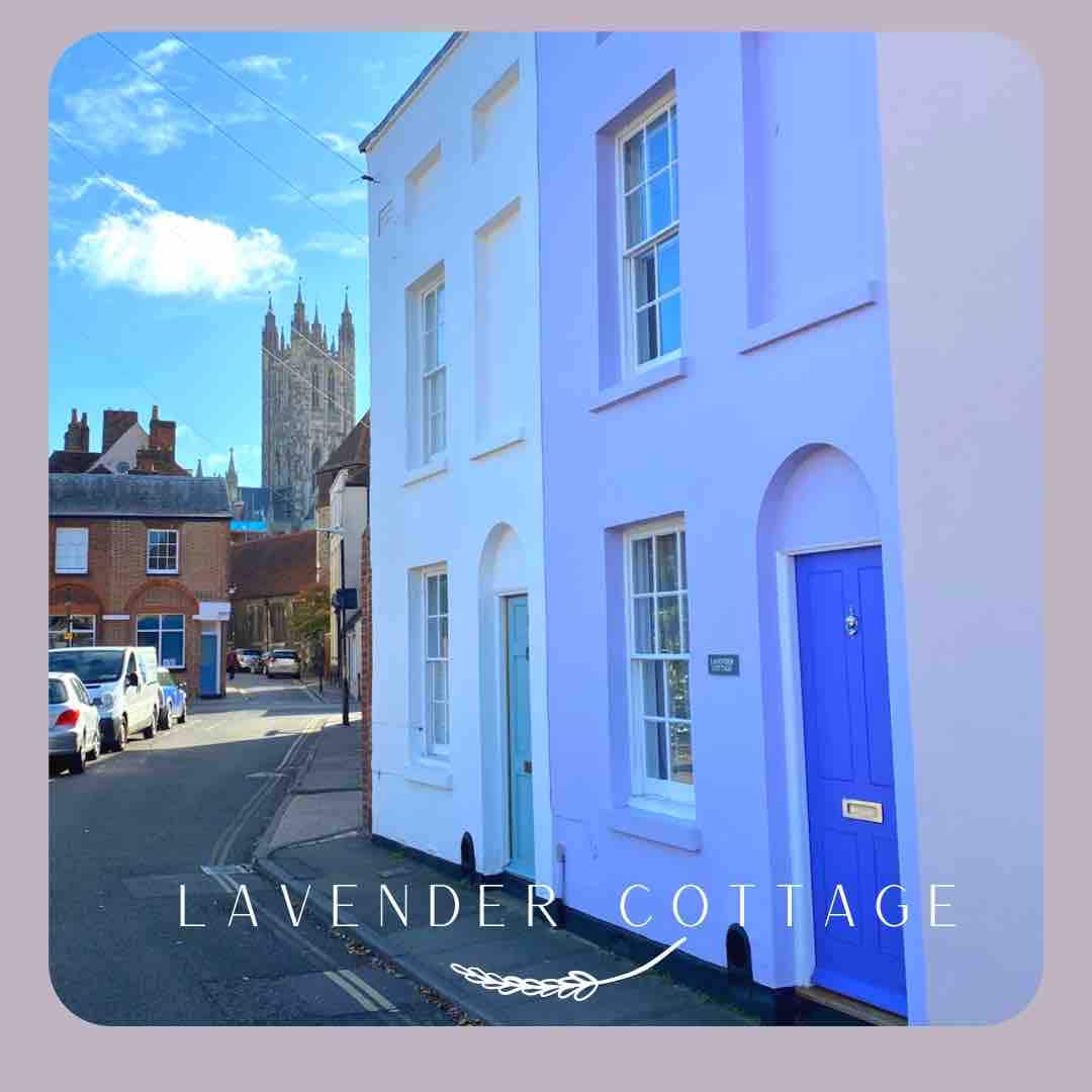 A charming lilac exterior of Lavender Cottage is presented alongside a blue door. The distant view of the Cathedral towers above the quaint street, showcasing a historic setting. The image captures the unique character of the neighborhood with residential buildings lining the street.