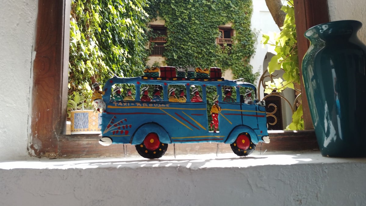 A colorful decorative bus figure is displayed on a windowsill. The vibrant blue bus features intricate details and is adorned with a luggage rack. Natural light filters through the window, highlighting the lush greenery outside and elements of the traditional architecture.