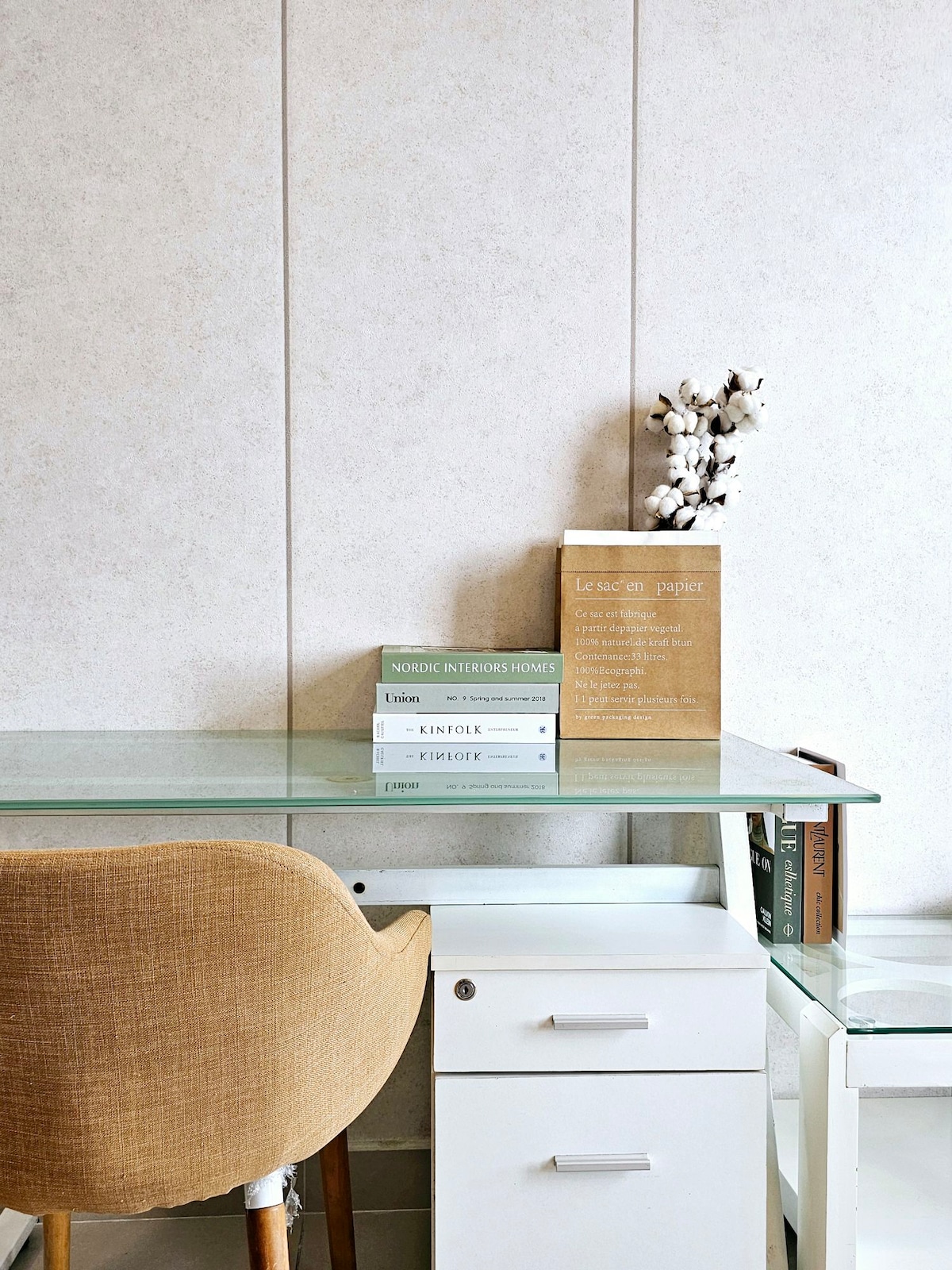 A modern workspace is presented, featuring a glass-topped desk and a comfortable chair. A stack of books is neatly arranged next to a decorative item, with light-colored walls providing a clean backdrop. A white cabinet is positioned beneath the desk, offering additional storage.