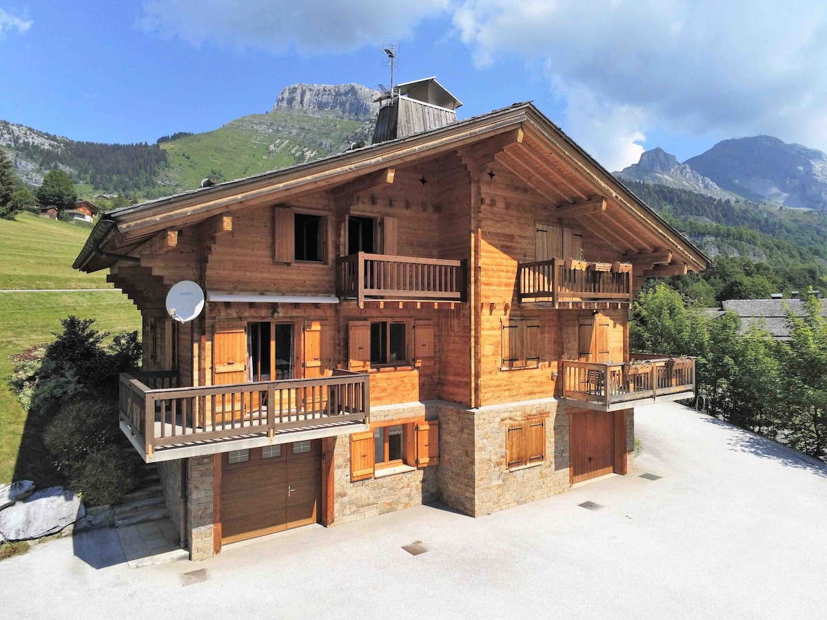 The chalet is crafted from natural wood, featuring multiple balconies and large windows that provide scenic views of the surrounding mountains. The stone base complements the wooden structure, which is set in a serene green landscape, enhancing the sense of tranquility.