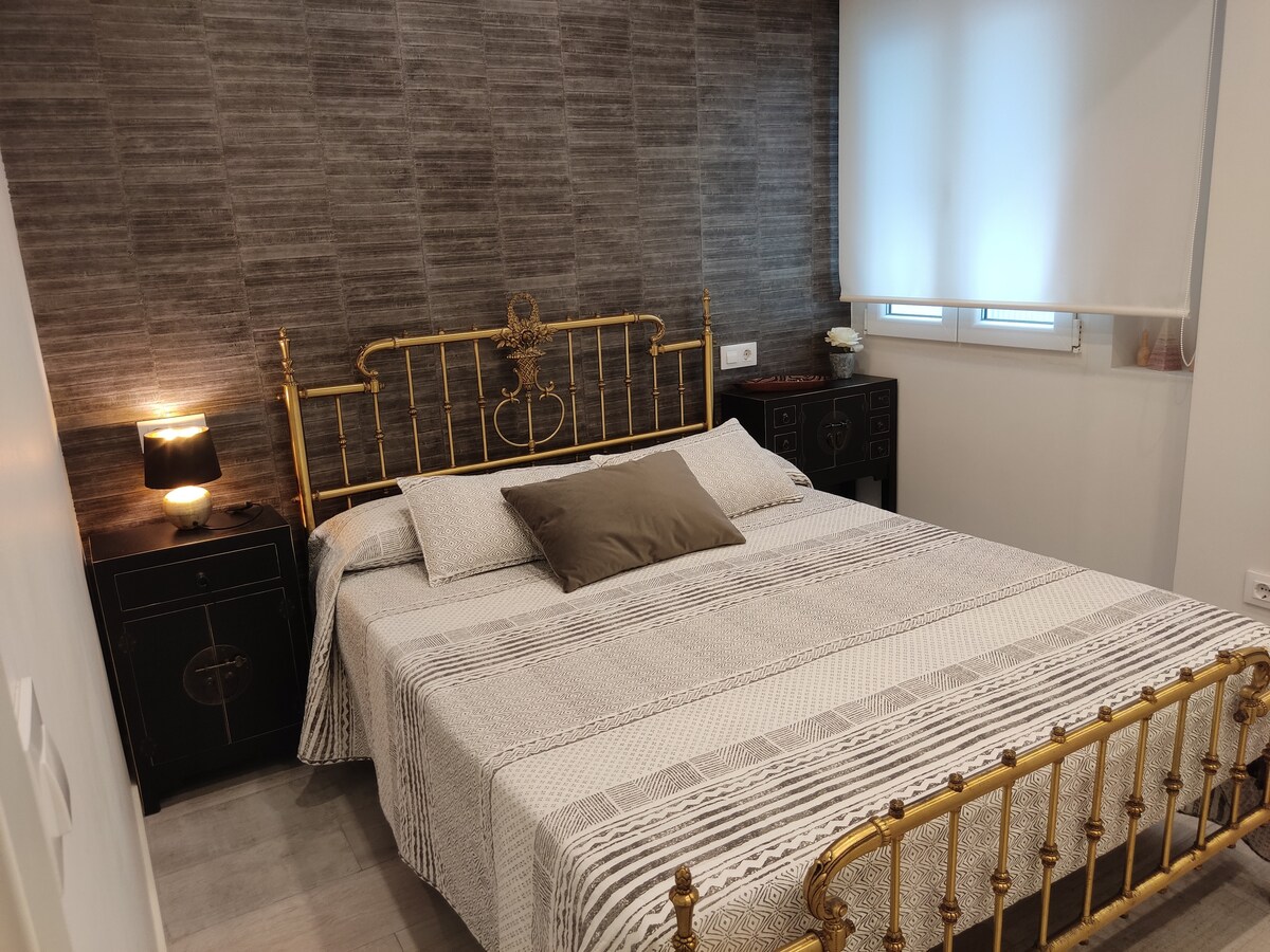 A cozy bedroom is highlighted by a gold metal bed frame, adorned with a neutral-toned blanket and a decorative pillow. Dark bedside tables with lamps provide subtle lighting, while a textured gray wall adds depth to the room's design.