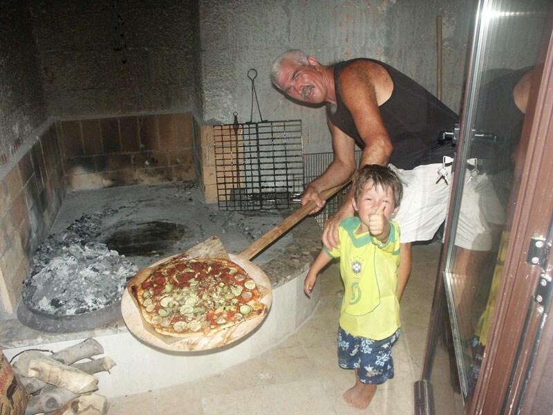 A traditional pizza oven is showcased, with a freshly made pizza being placed inside by an adult. A child stands nearby, observing the process. The rustic stone oven and wooden pizza peel are prominently featured, highlighting the art of pizza making.