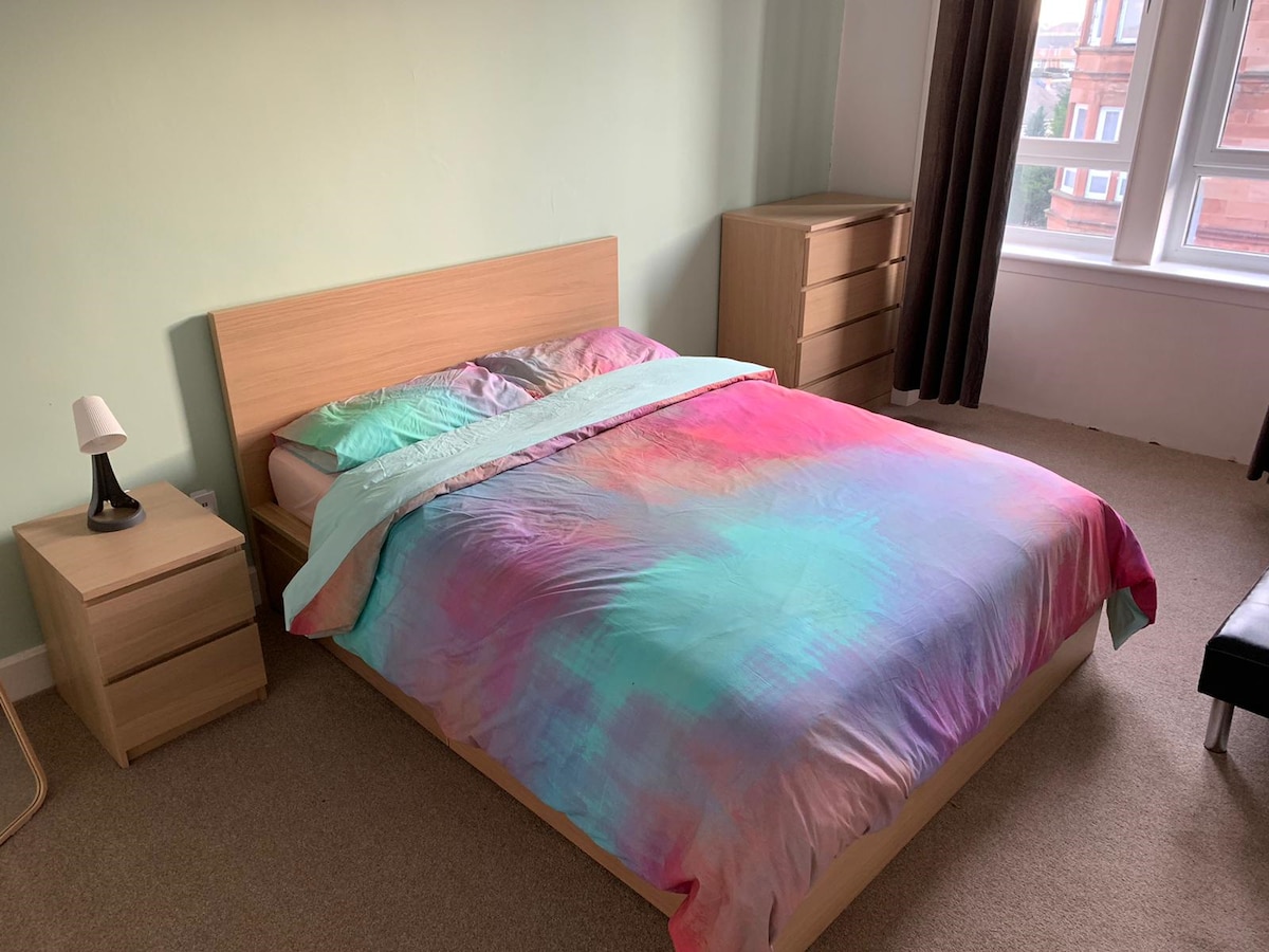 A spacious bedroom is presented, featuring a king size bed with a multicolored duvet cover. Light green walls provide a fresh backdrop, and a bedside table with a lamp is positioned next to the bed. A wooden dresser sits nearby, and large windows allow natural light to enter.