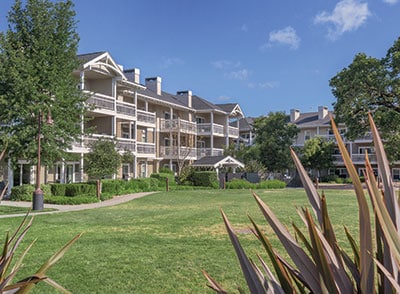 The exterior of a three-story building is visible, featuring multiple balconies and white railings. The building is surrounded by well-kept grass and shrubs, creating a green space. Blue skies and a few clouds complete the serene setting.