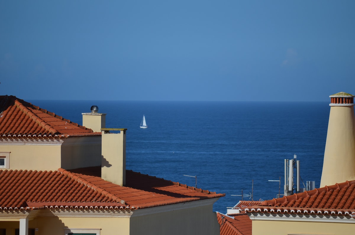 A distant sailboat glides across the expansive blue ocean, visible beyond the terracotta-tiled rooftops. Bright blue skies provide a clear backdrop, enhancing the serene coastal view.