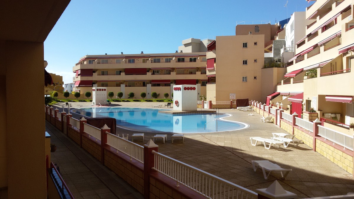A spacious outdoor area features a large swimming pool surrounded by sun loungers and neatly maintained grounds. The building exterior showcases a mix of beige and red tones with balconies, and the clear blue sky enhances the inviting atmosphere of the setting.