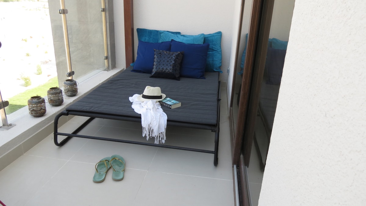 A cozy balcony area is furnished with a daybed adorned with blue pillows and a soft blanket. A straw hat, book, and white shawl rest casually on the bed. A pair of sandals is placed nearby, adding to the inviting atmosphere.