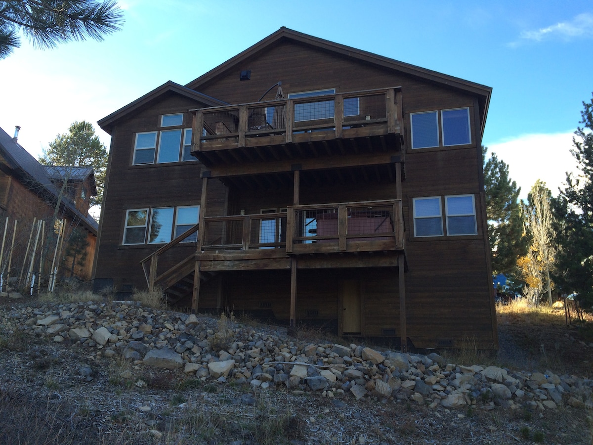The exterior of a multi-story mountain retreat showcases a warm wooden facade. Two large decks are visible on the upper level, providing outdoor space. The property is situated on a gently sloping lot with natural landscaping featuring rocks and pine trees.