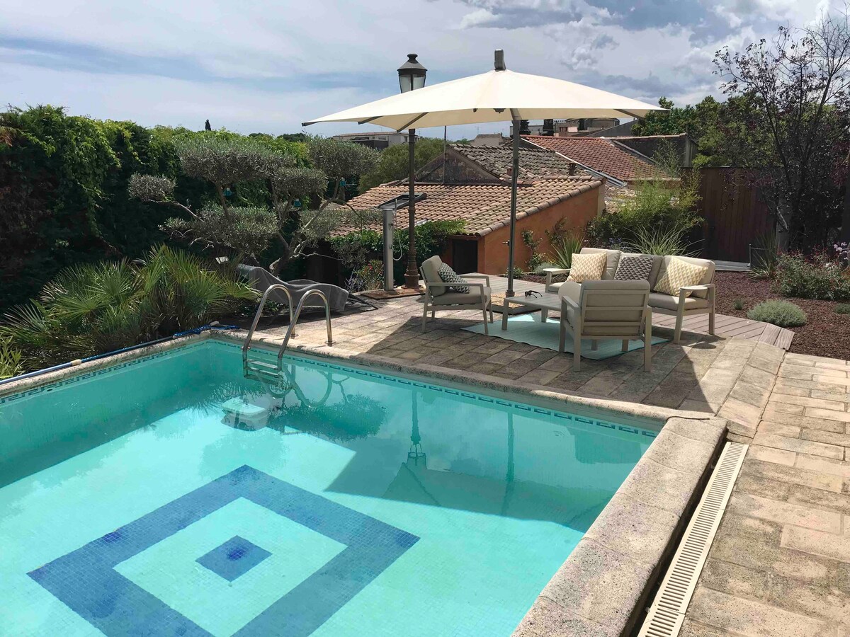 A private swimming pool is surrounded by tropical plants and outdoor seating. Light-colored lounge chairs and a dining table sit under a large umbrella, offering a shaded area. The pool features a geometric design at the bottom, with stone tiles lining the pool deck.