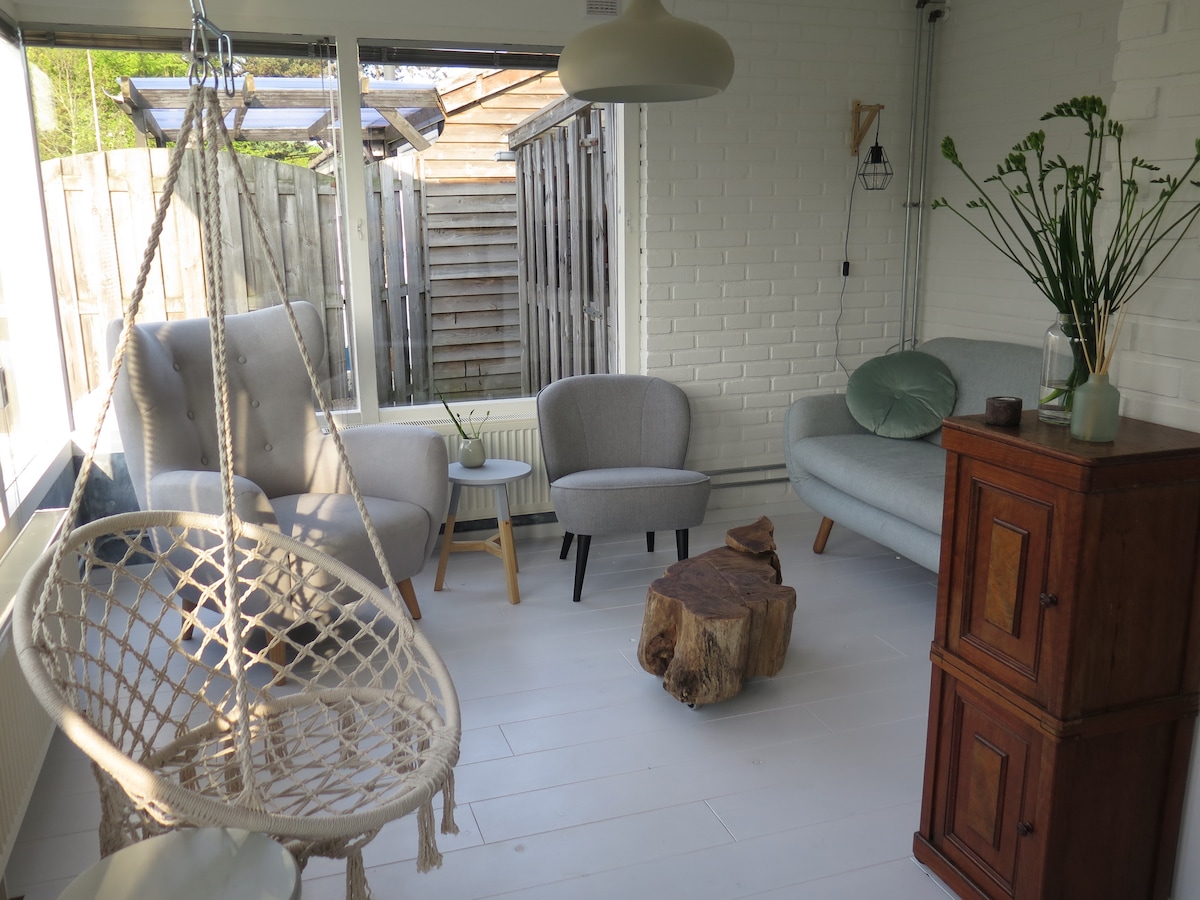 A bright and airy seating area features a hanging chair and two upholstered chairs complemented by a small wooden table. Light streams in through large windows, and a wooden cabinet adds character to the space. The walls are adorned with a crisp white finish.