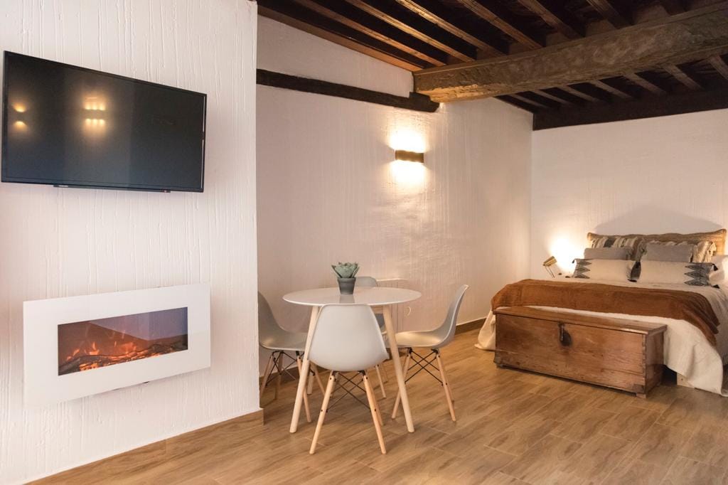 A cozy area features a spacious bed with a neutral-toned comforter and accent pillows. A circular dining table is surrounded by four modern chairs. A wall-mounted TV is positioned above a sleek, electric fireplace, creating a relaxed ambiance in the well-lit space.