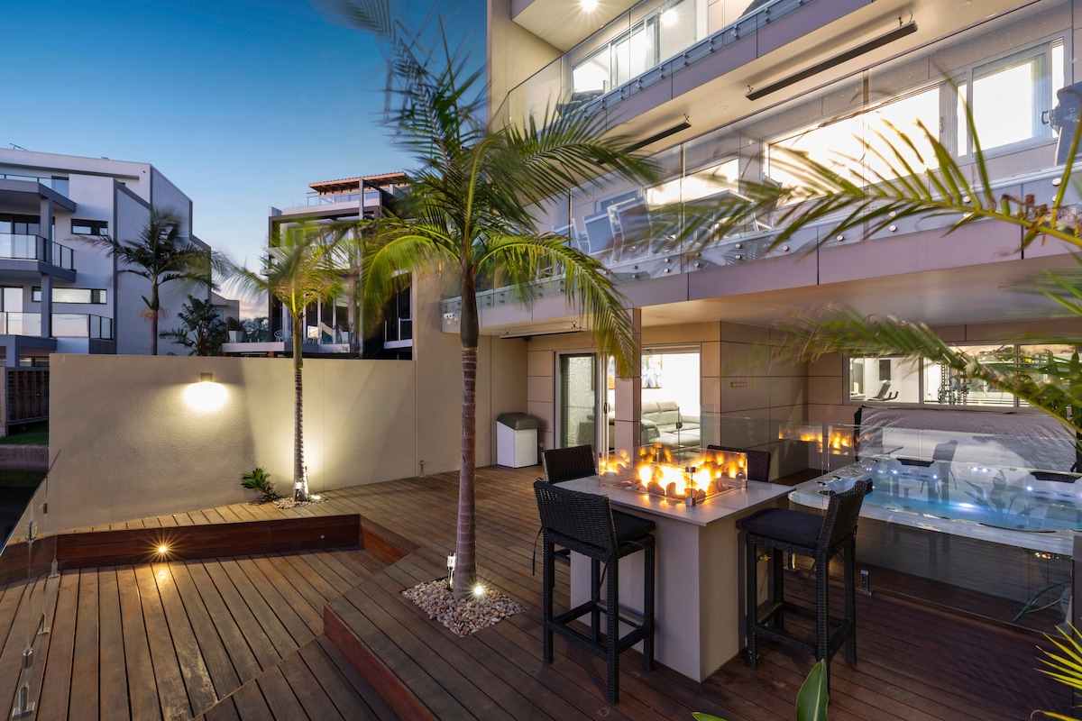 A spacious outdoor area is highlighted by a wooden deck featuring a dining table with chairs. Subtle ambient lighting, applied to the wall, enhances the space. Nearby, a heated spa is visible, surrounded by lush palm trees, creating a serene and inviting environment.