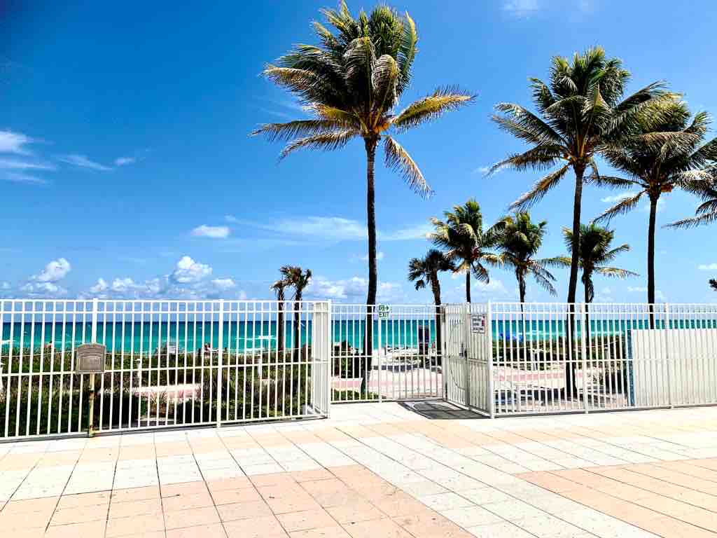 A view through a gated entrance reveals a vibrant beach scene. Palm trees sway gently against a clear blue sky, leading to the turquoise waters of the ocean in the background. A paved walkway runs beneath the gate, connecting to the beach area.