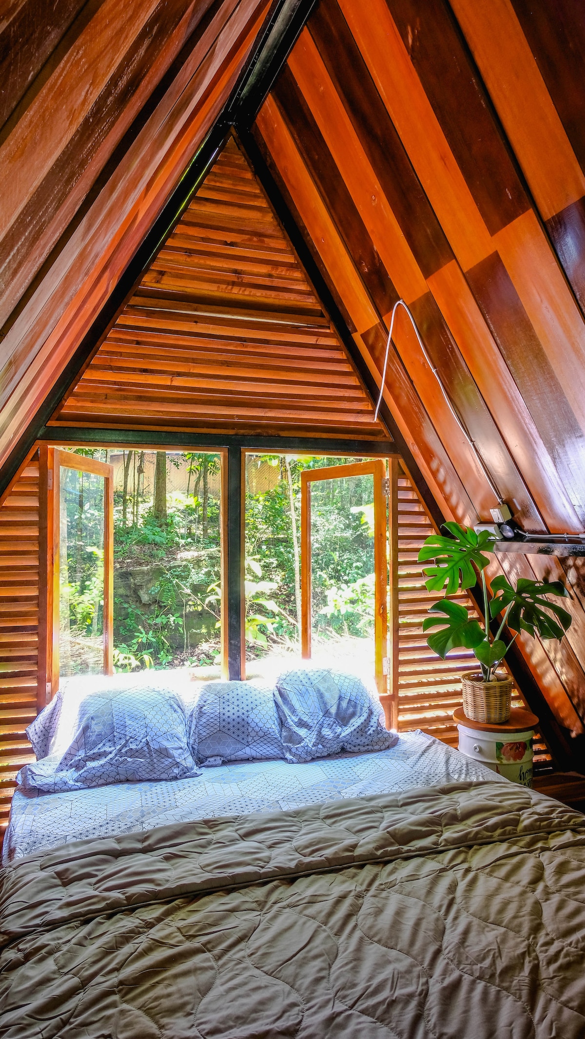A cozy loft bedroom features a spacious bed adorned with a light-coloured blanket. Large windows framed in wood offer views of the surrounding greenery, allowing natural light to illuminate the room. Potted plants add a touch of nature to the inviting space.
